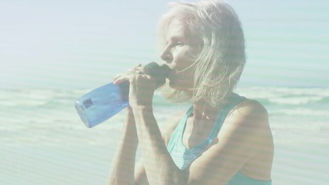 Feeling thirsty, senior woman lifting blue bottle and drinking at shoreline, showing health