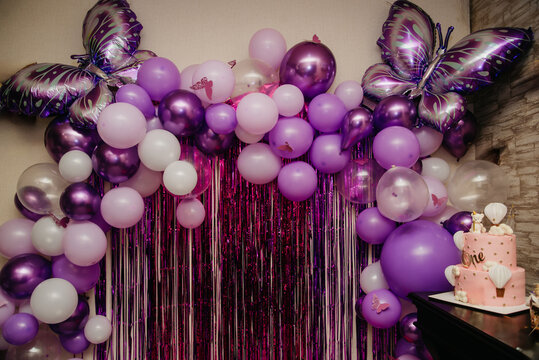 A photo zone made of purple balloons with butterflies attached to them and shiny ribbons is created for a birthday celebration, with a pink cake next to it