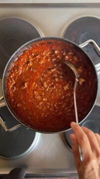 overhead view of person preparing meal, holding ladle stirring freshly cooked bolognese sauce cooking in large saucepan on electric stove top at home in kitchen