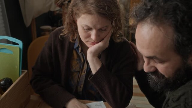 Close up shot of bearded man supporting his stressed wife, sitting together at table with past due bills in poor apartment
