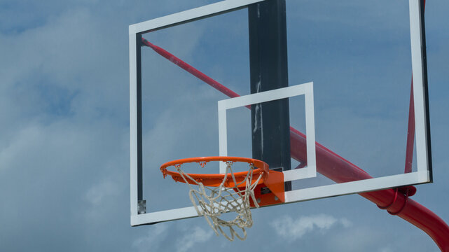 Outdoor basketball hoop with orange rim and transparent backboard against cloudy sky