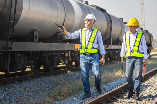 Railway engineers using walkie talkies while inspecting oil tank wagons on tracks, Asian railway foreman workers in safety vests and hardhats discussing maintenance near a freight train