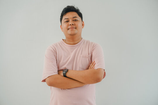 a man wearing a pink t-shirt and a watch stands with his arms crossed. he looks confident and serious in a plain gray setting.