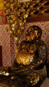 The dark-colored statue is heavily adorned with numerous small, square pieces of gold leaf, particularly prominent on its large belly and face
