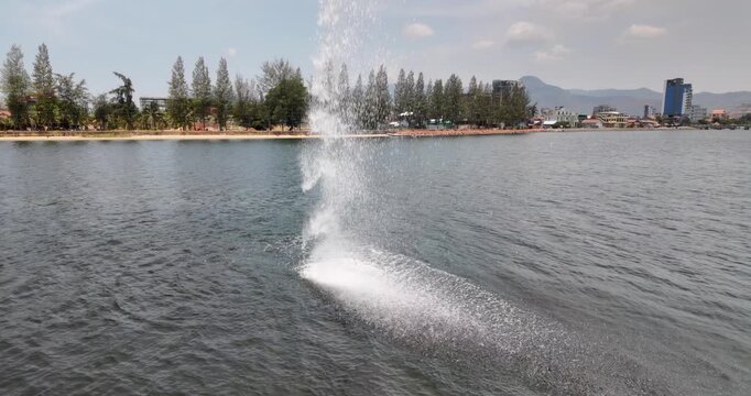 Slow motion of a large plume of water spraying from the Seahorse statue into the river in Kampot with a tree-lined shore and distant mountains in the background.