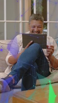 Vertical video: Mature man sitting sofa tapping tablet causing blue teal tech overlay for leisure