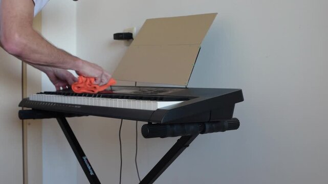 man cleaning synthesizer keys indoors meticulous hands wipe dust from black digital keyboard on collapsible stand, orange microfiber cloth, open music stand, cables dangling,