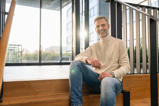 Smiling man in casual clothing sitting on stairs in a modern office