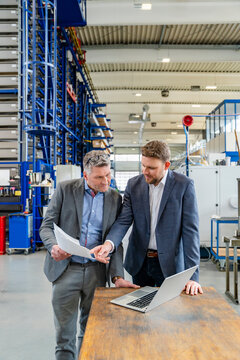 Managers discussing document with laptop in production hall