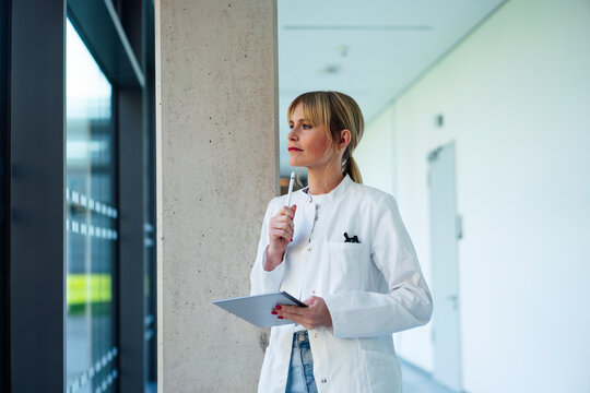 Thoughtful female doctor standing with tablet PC and digitized pen looking out through window in hospital