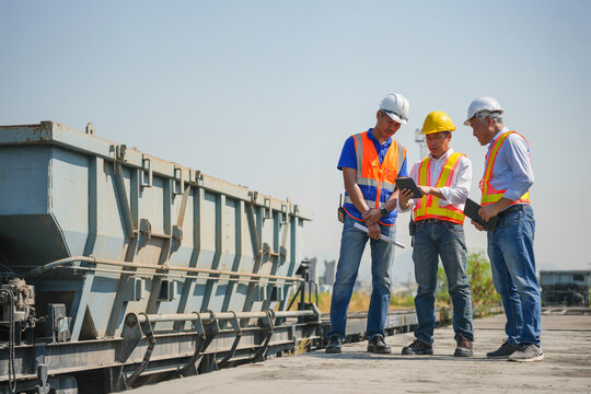 Team of railway engineers using digital tablet for freight train inspection and logistics planning, Group of inspectors checking maintenance schedule on tablet near large industrial freight wagon