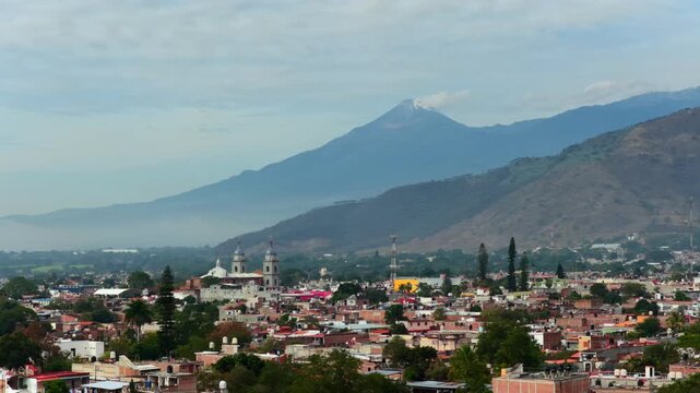 Tuxpan cityscape with low rise buildings and church towers spread across the valley, backed by the Colima Volcano mountain range with light snow on the summit in Jalisco