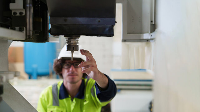 Male worker in yellow safety vest meticulously inspects CNC machine spindle with tablet, performing precision maintenance and quality control in modern factory environment.