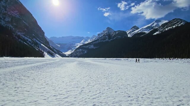 People walk across frozen Lake Louise in Banff National Park, surrounded by snow-covered mountains, dense evergreen forest, bright daylight, and dramatic Canadian Rockies scenery.