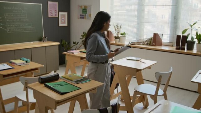 Full lockdown shot of young female teacher handing out health education textbooks on student desks during break while getting ready for lesson in middle school, human body diagram on chalkboard