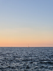 Fototapeta premium Calm sea at sunset with soft pastel sky and distant sailboats on the horizon, minimal seascape background with copy space.