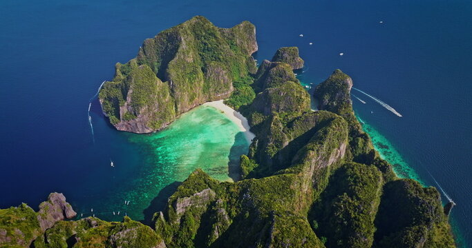 Phi Phi Island Maya Bay aerial: towering limestone cliffs draped in lush green, pristine white sand and vibrant turquoise water with boats cruising coast. Summer vacation paradise, travel to Thailand