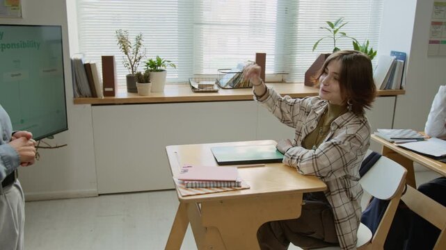 Medium pan shot of embarrassed, giggling teenage Caucasian girl raising hand to ask question to teacher during lesson on pregnancy, concerning prevention, conception and reproductive health