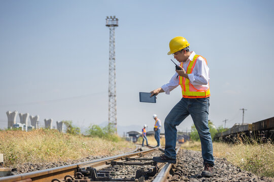 Railway engineer monitoring train tracks with digital tablet and radio communication at rail yard, Professional Asian inspector and team in safety vest and helmet working on railway infrastructure