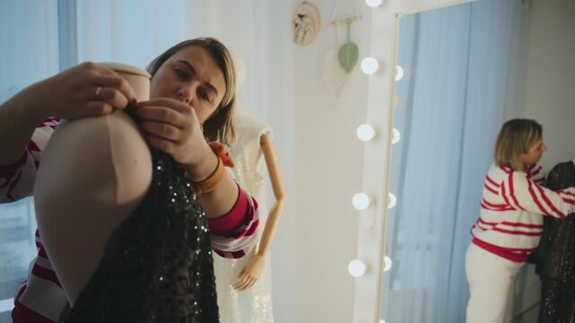Woman tries on sparkly dress in dressing room mirror. She adjusts garment with focused attention. Bright lights surround mirror reflecting her actions. Suitable for fashion, retail