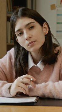 Vertical medium portrait of demure teenage Caucasian girl in pink jumper sitting at desk, looking at window then turning to camera, while writing trimester test on health education course