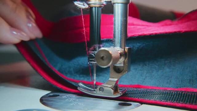 Close-up of hands guiding fabric under sewing machine needle. Thread pierces material with precision. Fabric shows red and dark blue stripes. Hands steady, focused on stitching task