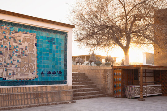 Ceramic tile map of Ichan Kala monuments and camel caravan sculpture at sunset in Khiva. Golden hour light illuminates the historic old town entrance with a souvenir stall and bare winter tree. Ide...