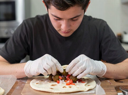 A young male chef wearing gloves while adding toppings to a pizza base at an industrial kitchen counter.