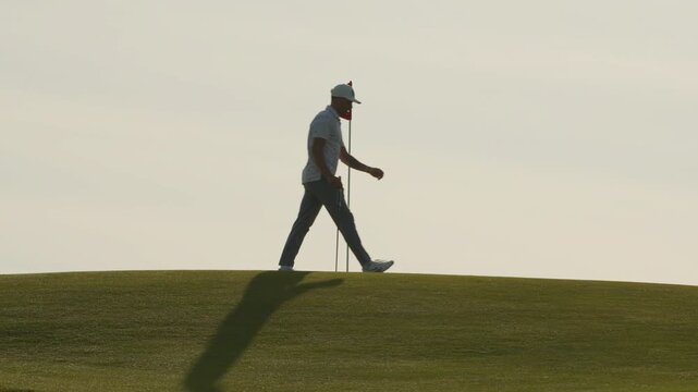 Male Golfer Walks on Green in Slow Motion, Flag Waving in Wing, Sunset