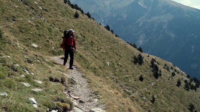 Static FHD shot of a backpacker walking along a rocky alpine path on a steep grassy slope with dramatic Pyrenees mountains in the background.