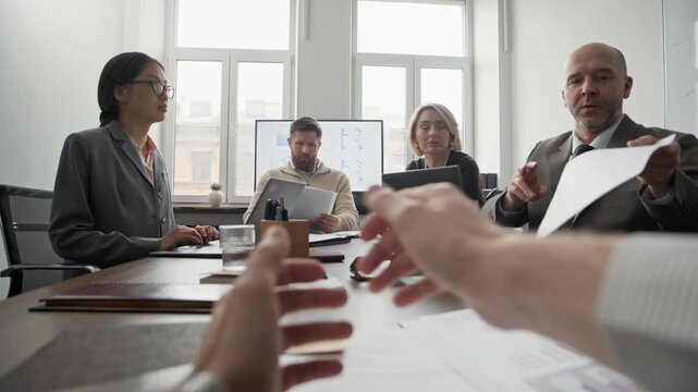 POV of anonymous man sitting at desk with his colleagues during office meeting and giving paper with diagrams to man