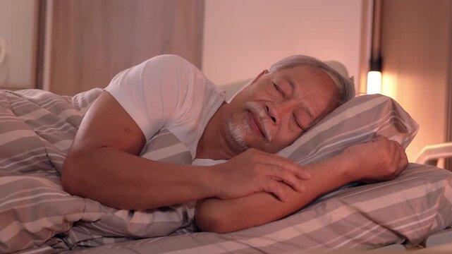 Happy elderly senior man smiling while sleeping peaceful in comfortable bed resting head on soft striped pillow enjoying quiet restful slumber inside cozy domestic bedroom during calm night
