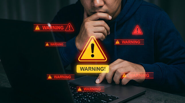 Man in a dark hoodie, deep in thought while working on a laptop showing multiple warning symbols and alerts.