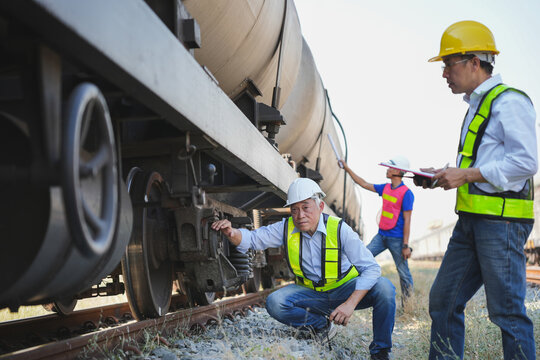 Team of Asian engineers in safety vests inspecting wheels and undercarriage of oil tank train, Professional railway technicians performing maintenance check on industrial freight wagon at rail yard