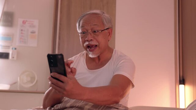 Happy senior asian man wearing glasses and white undershirt holds smartphone in bed room at night expressing excited joyful smile during online video call experiencing modern technology