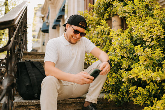 Caucasian Man On Steps Checking Phone, Relaxed Freelancer Wearing Cap And Sunglasses, Backpack Beside Him,