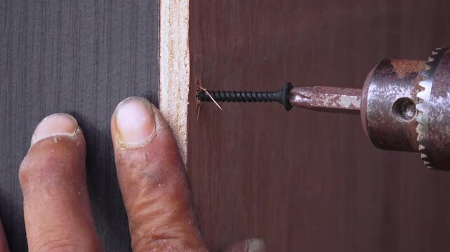 A close-up showing a finger positioning a black screw against the edge of a plywood panel before using an electric drill.