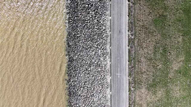 A static top-down drone shot of the road atop the Barbate dam, creating a symmetrical contrast between the water, rock reinforcement, and land.