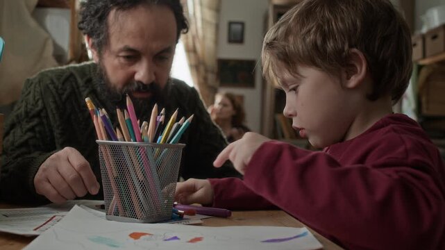 Low angle shot of bearded man drawing with colored pencils with his young son in small poor apartment