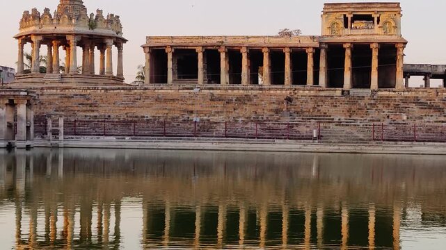 Stone pavilion, steps and Bhuvaneshwari Mantapa Dome reflected in still water at a historic Melukote Kalyani pond during sunset