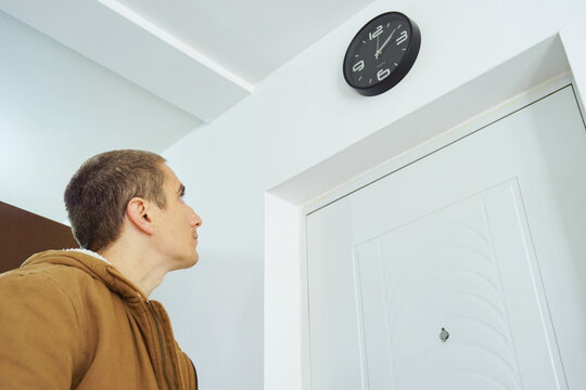 Man looking up at wall clock beside apartment entrance door before leaving home. Conveys departure timing awareness, schedule control, punctuality pressure, and personal time management discipline.