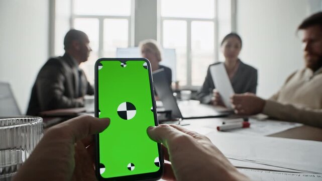 POV of unrecognizable man holding smartphone with neon screen while sitting at desk during office meeting