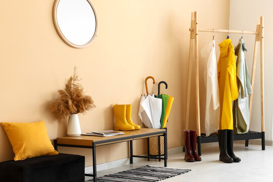 Interior of room with bench, gumboots, umbrellas and different colorful raincoats hanging on clothes rack near beige wall