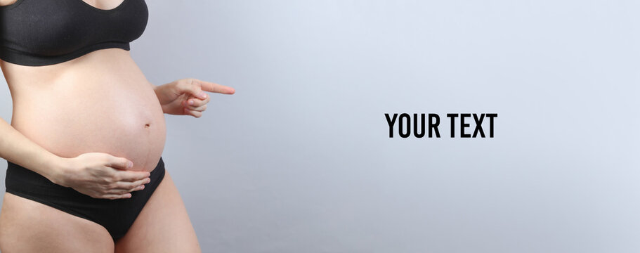 Pregnant woman in underwear points with a finger on copy space, gray background. Copy space for your text
