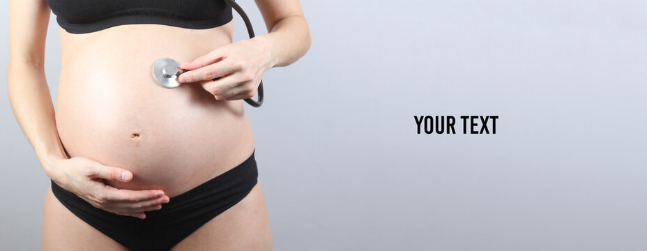 Pregnant woman listening to her belly with stethoscope on gray background. Copy space for your text