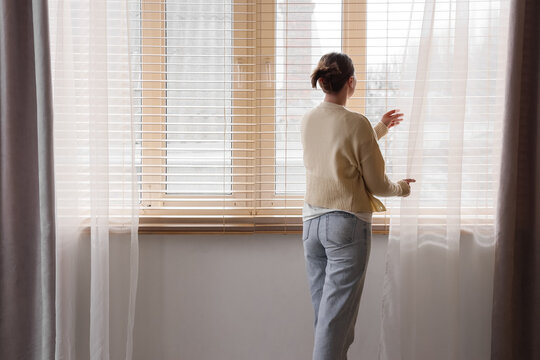 Young woman opening white curtains at home, back view