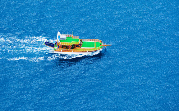 Aerial view of traditional Turkish wooden boat with green top, known as gulet, sailing on vibrant blue Mediterranean sea water. Vessel leaves white wake trail. Travel and tourism concept