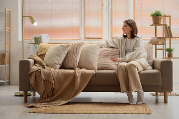 Obraz premium Beautiful young woman sitting on cozy sofa with soft pillows in living room