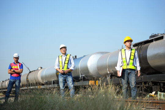 Group of Asian engineers in safety vests and hardhats standing by oil tank train at rail yard, Professional team of railway technicians and logistics managers supervising freight train operations