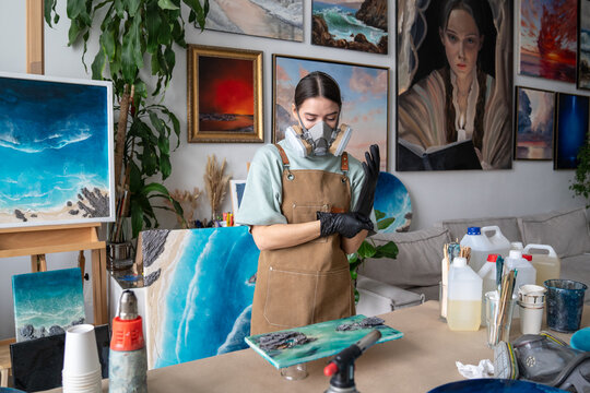 Serious woman artist preparing for work with epoxy resin in workshop, wearing gloves and respirator for safety, focused female crafter uses special equipment and tools for liquid painting in studio.